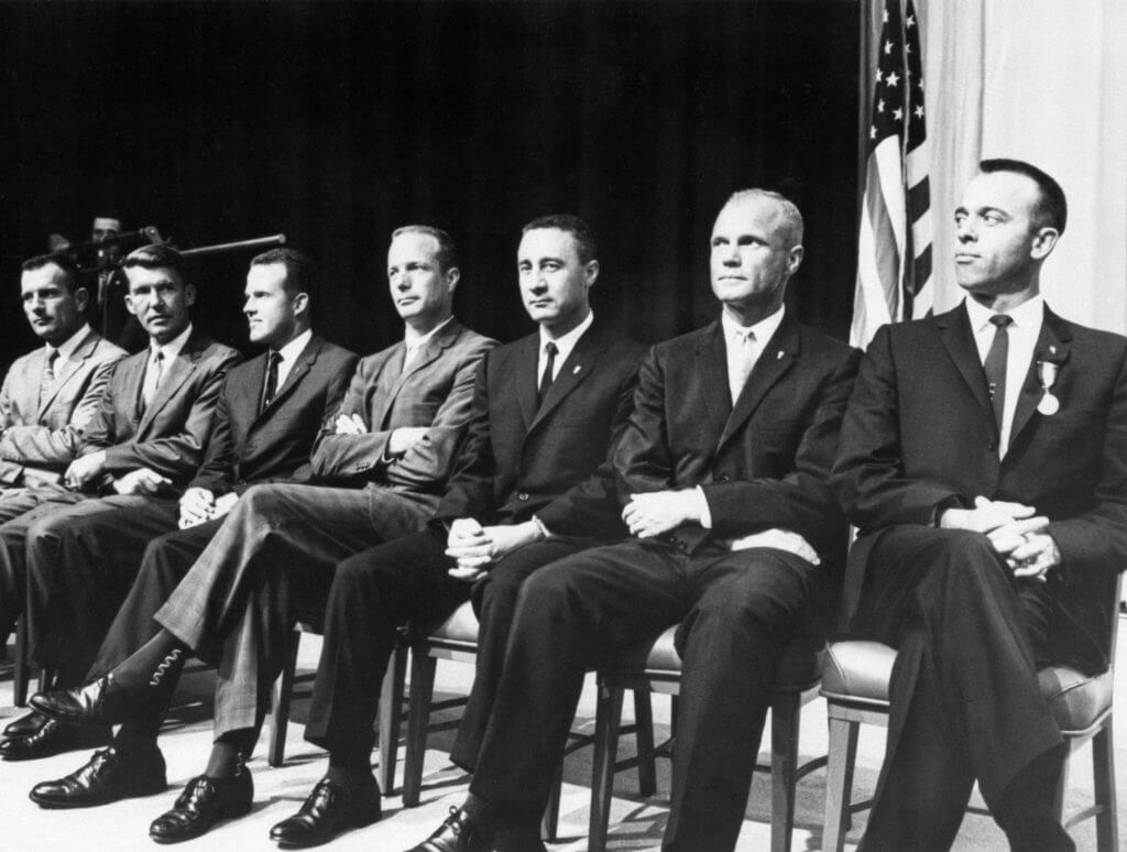 Grupa astronautów znana pod nazwą Mercury Seven, siedzących na krzesłac. Od lewej:  Donald K. Slayton, Walter M. Schirra, Jr., L. Gordon Cooper, Jr., M. Scott Carpenter, Virgil I. Grissom, John H. Glenn, Jr. and Alan B. Shepard, Jr.  : 